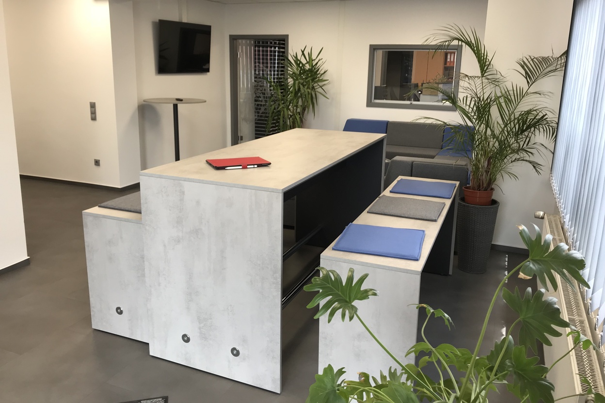 Einladender Büroloungebereich mit grauem Sofa, Pflanzen und einem hohen Tisch mit rotem Buch und blauen Matten.