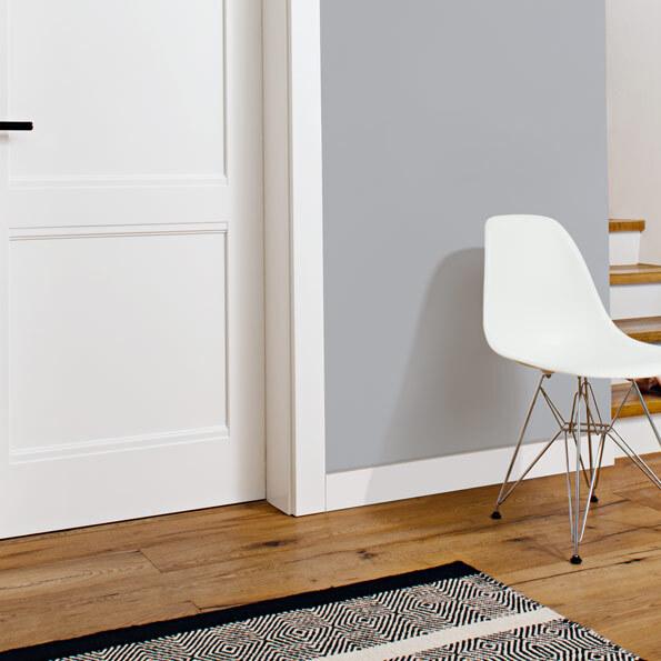 Stilvolle weiße Tür nach Maß mit klassischem Kassettendesign und Edelstahlgriff in einem modernen Wohnraum. Der Raum ist mit grauer Wandfarbe, Holzfußboden und einem weißen Designstuhl gestaltet. Eine Treppe mit Holzstufen f&u