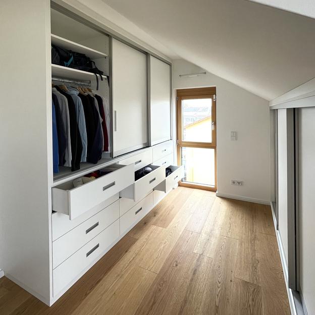 Ein weißer, maßgeschneiderter Schrank, der nahtlos unter einer Dachschräge integriert ist, mit Schubladen und Hängeregistern, der den verfügbaren Raum effizient nutzt.
