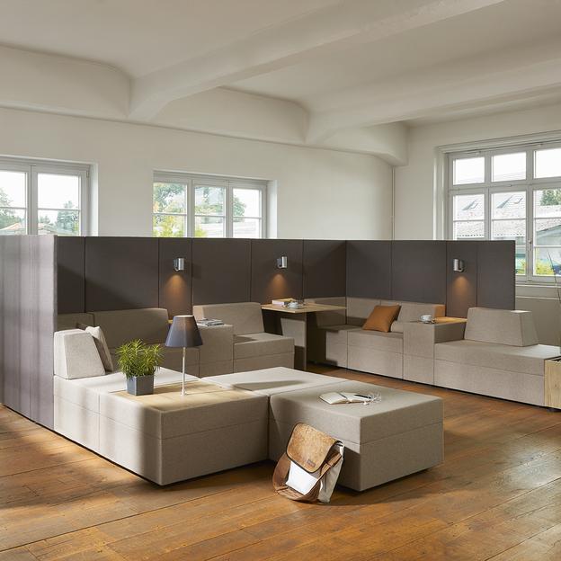 Elegante Bürolounge mit modularen Sitzmöbeln Ein modern gestaltetes Büro mit modularen Sitzmöbeln in Beige und Braun, aufgeteilt durch dunkle Trennwände, beleuchtet durch schlichte Lampen. Ein großer Holzboden und hohe Fenster runden den Raum ab.