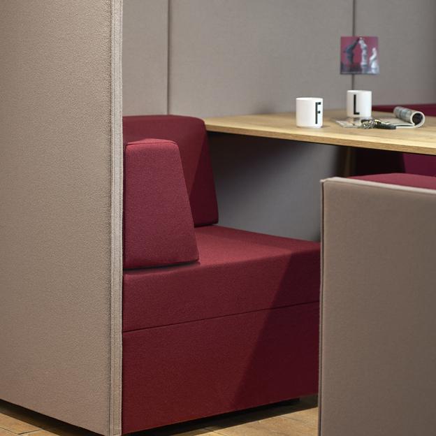 Arbeitsplatz mit bordeauxrotem Sitzmöbel Detailansicht eines Arbeitsplatzes mit einem bordeauxroten Sitzmöbel, flankiert von grauen Trennwänden und einem Holztisch mit persönlichen Gegenständen.
