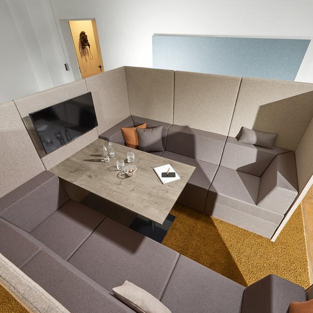 Innovatives Sitzmöbel-Design in modernem Arbeitsraum Ein Blick von oben auf eine innovative Sitzmöbeleinheit in Beige und Braun mit integriertem Tisch, umgeben von einer lebendigen Holzbodenfläche und einer künstlerischen Wanddekoration.