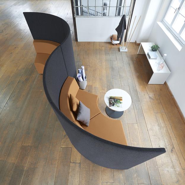 Geschwungenes Sitzmöbel in einem offenen Loft Luftaufnahme eines geschwungenen Sitzmöbels in Dunkelgrau und Karamell, platziert auf einem rustikalen Holzboden in einem hellen, offenen Loft mit großen Fenstern.