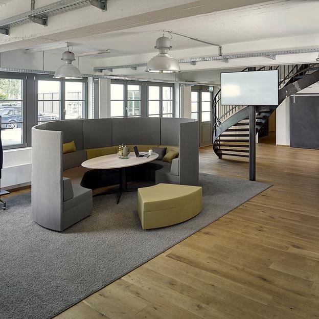Modernes Büro mit stilvollen Sitzmöbeln und Loft-Design Ein stilvoll eingerichtetes Büro mit modernen Sitzmöbeln in Grau und Limettengrün, arrangiert auf einem grauen Teppich über einem Holzboden, mit einer eleganten Wendeltreppe im Hintergrund.