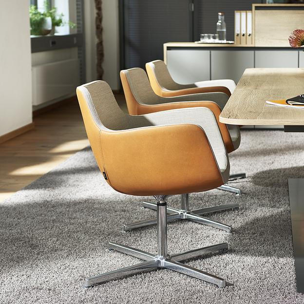 Stilvolle Bürostühle in einem modernen Büro Eine Reihe eleganter Sitzmöbel in Orange und Grau, angeordnet um einen Holztisch in einem sonnendurchfluteten Büro, ideal für Meetings oder Pausen.