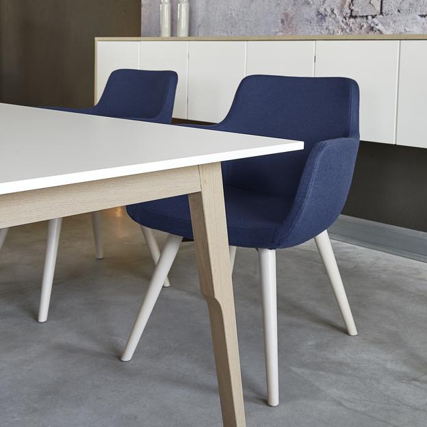 Elegante blaue Stühle an einem modernen Esstisch Zwei dunkelblaue Stühle als Sitzmöbel neben einem weißen Tisch mit Holzbeinen, positioniert in einem stilvollen Raum mit dekorativer Wand und minimalistischem Schrank.
