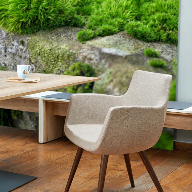 Moderner Stuhl in einem naturnahen Arbeitsbereich Ein elegantes Sitzmöbel mit beige-brauner Polsterung und dunklen Holzbeinen, platziert neben einem hölzernen Schreibtisch mit Blick auf üppiges Grün außerhalb des Fensters.