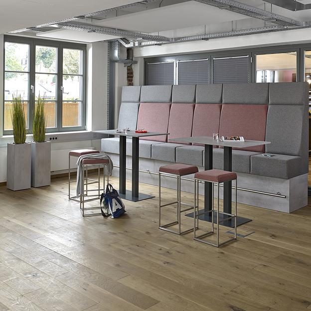 Stilvolle Sitzmöbel in einem modernen Büro-Café Ein Büro-Café mit einer großen, mehrfarbigen Sitzbank und Barhockern an einem hohen Tisch, umgeben von natürlichem Holzboden und großen Fenstern.