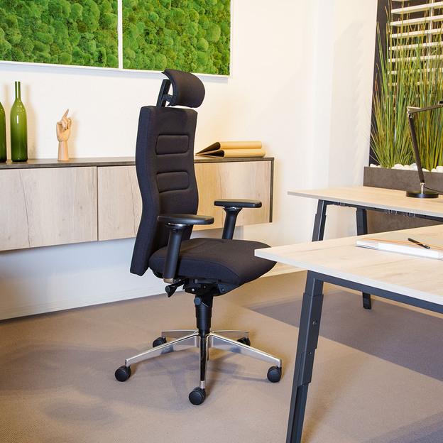 Ergonomischer Bürostuhl in modernem Büro Ein modernes Büro mit einem ergonomischen schwarzen Sitzmöbel, flankiert von einem hellen Holztisch und dekorativen Elementen wie grünen Flaschen und Wandbildern mit Moos.