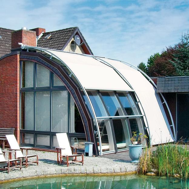 Modernes Haus mit einer einzigartigen gewölbten Struktur als Sonnenschutz über einer Glasfassade, die auf einen Garten und einen Teich blickt. Lounge-Stühle stehen im Außenbereich bereit.